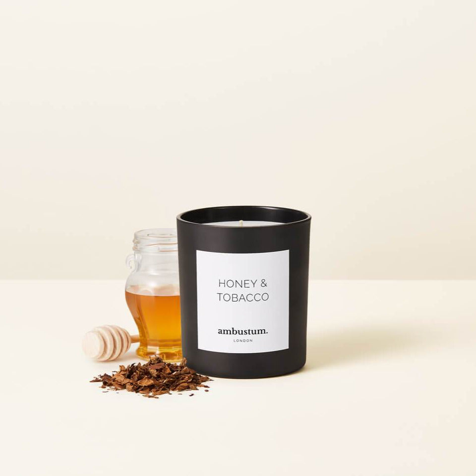 A photograph of the Honey & Tobacco candle with a honey pot layered behind, the candle in the black glass packaging 