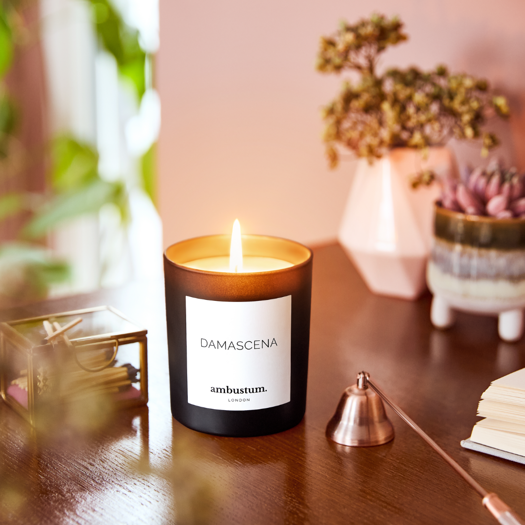An ambustum Damascena candle in a glass candle jar with elegant white labelling. Blending sweet and fruity notes of blackcurrant, damson plum, and mandarin with delicate floral scents of carnation, rose, lily, and geranium.
