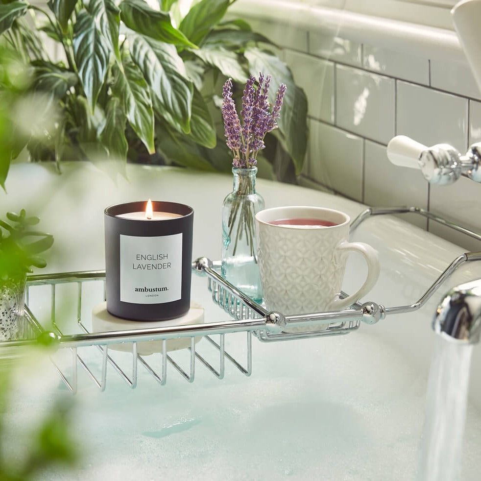 A lit lavender-scented candle, a cup of tea, and a small vase with lavender sprigs on a bathtub caddy surrounded by green plants