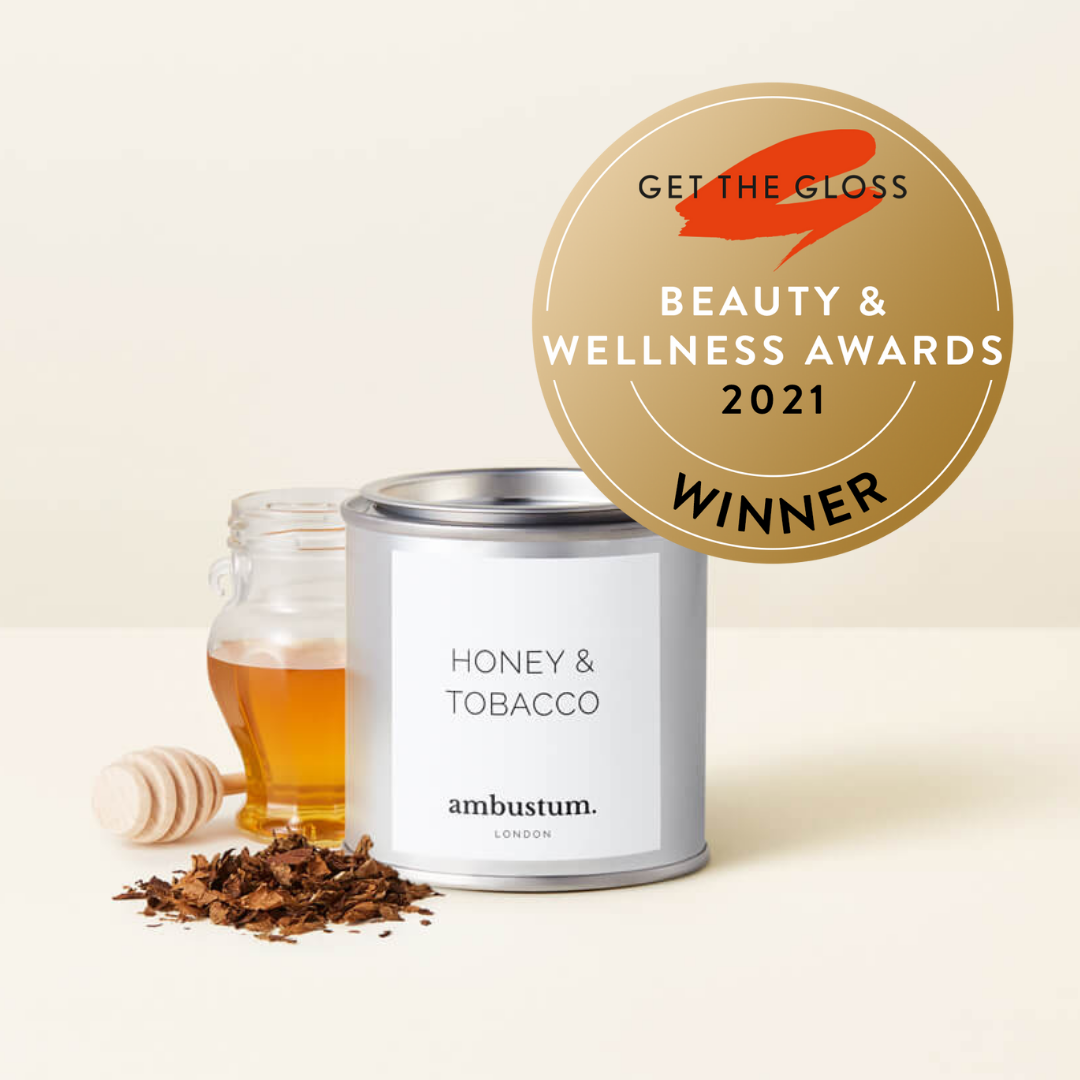 An ambustum honey and tobacco candle in a tin with elegant white labelling. The scent profile is A dark and powerful blend of tobacco leaf, softened with notes of honey, amber and patchouli. Perfect for creating a warm and inviting atmosphere.
