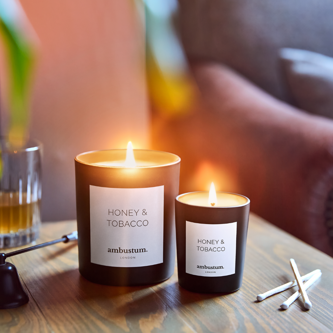 An ambustum honey and tobacco candle in a glass candle jar with elegant white labelling. The scent profile is a dark and powerful blend of tobacco leaf, softened with notes of honey, amber and patchouli. Perfect for creating a warm and inviting atmosphere.